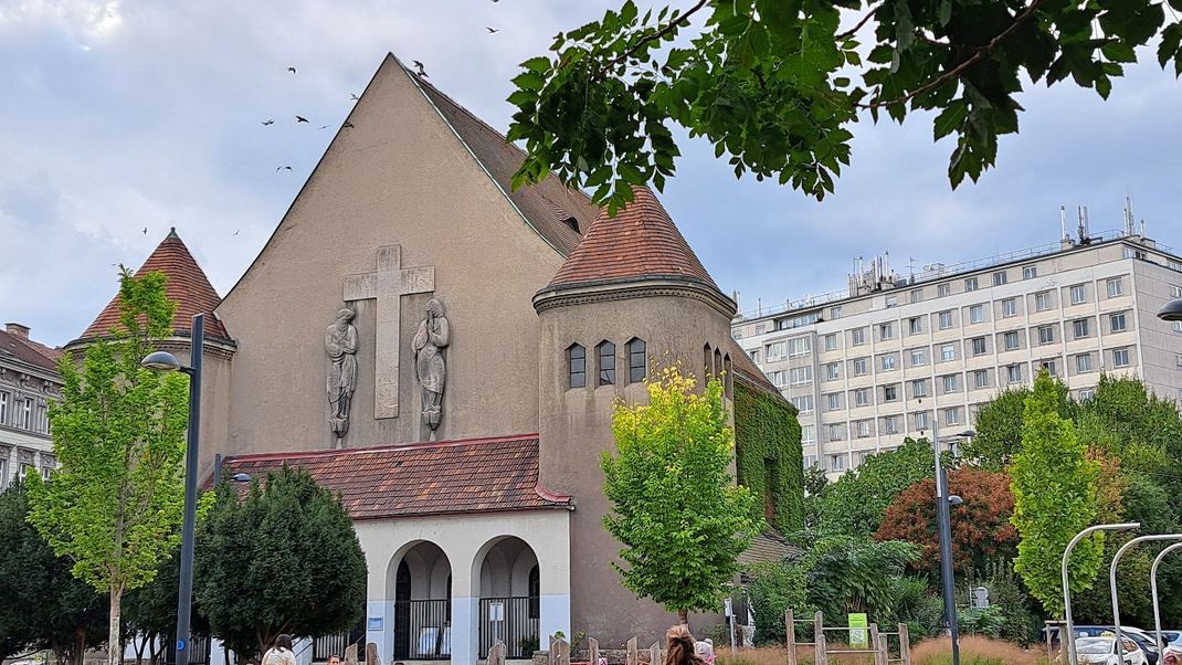 The slightly weathered main façade of the Protestant church, with a gable roof and a pointed gable. A cross and two figures in bas-relief are incorporated into the flat main façade. On either side are low towers with pointed hipped roofs. Between them lies the porch with round arches. In front of and to the sides are green tree tops; in the background stands a modern high-rise residential block.