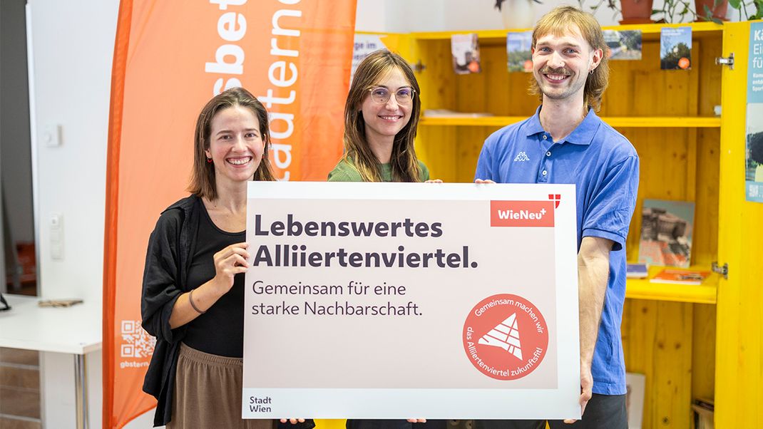 Three smiling young people looking at the camera, holding a large information board bearing the words ‘The Alliiertenviertel – a great place to live, working together for a strong neighbourhood’. In the background, a yellow display stand and an orange banner.