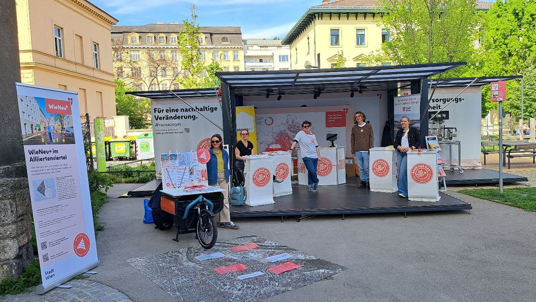 An open information stand with several people standing at display tables and information boards, with an urban landscape featuring Gründerzeit-era buildings and trees in the background.