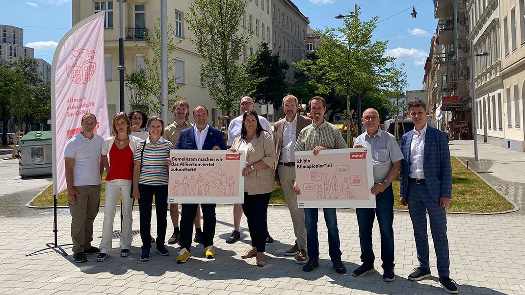 The photograph shows twelve people standing in a row in a square, smiling warmly at the camera. They are holding two signs with writing on them; on the left is a pink beach flag. In the background is a street lined with Wilhelminian-style houses and trees.