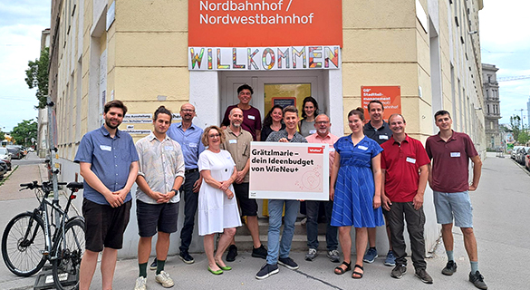 Group photo with Stephan Hartmann in the centre, holding a large information board. In the background is a Wilhelminian-style building and an entrance with the inscription ‘Norbahnhof/Nordwestbahnhof’ and ‘Welcome’ in colourful lettering.