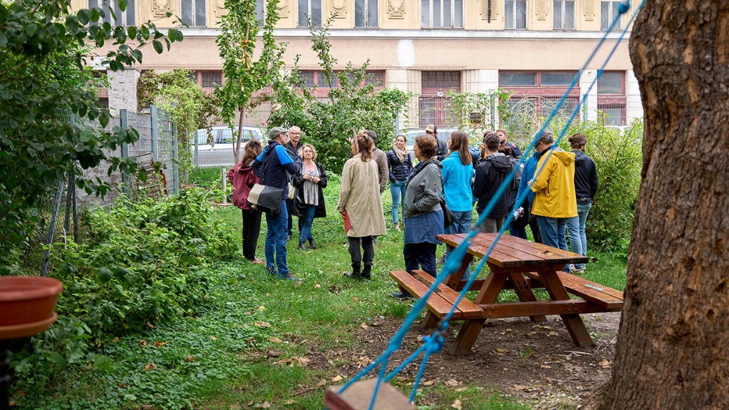A group of people standing in a wide circle in a garden, with a table and benches in front of them and, in the foreground on the right, a tree with a children’s swing. In the background is a historic old building.
