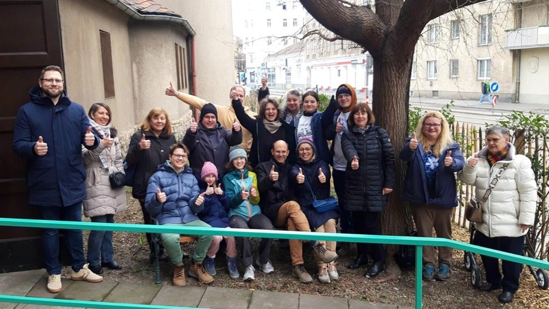 A cheerful group of people, facing the camera and giving a thumbs-up. Parts of the church building are visible on the left; on the right is a tree, with the street and buildings in the background.