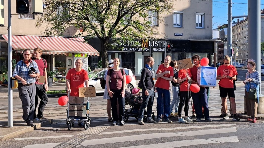 A group of people on a traffic island, some wearing red T-shirts, who are clearly waiting for the lights to turn green. Some are holding signs, others red balloons.