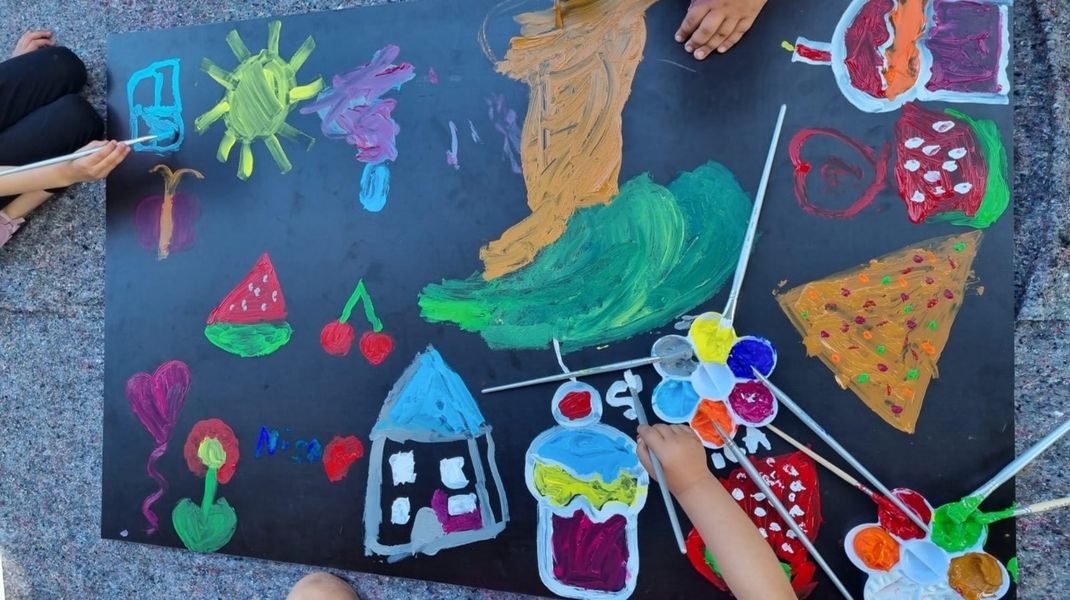 A black rectangular board is being painted in bright colours with children’s motifs by children’s hands. A paintbrush and a flower-shaped paint palette are also lying on the board.