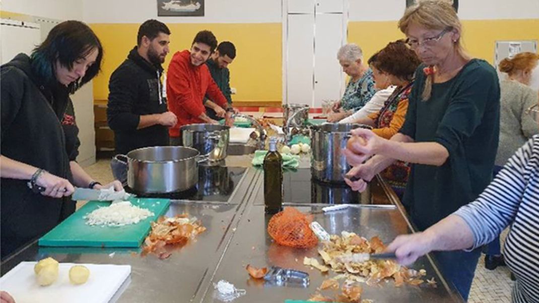 A group of young people are cooking together with a group of older women and men.