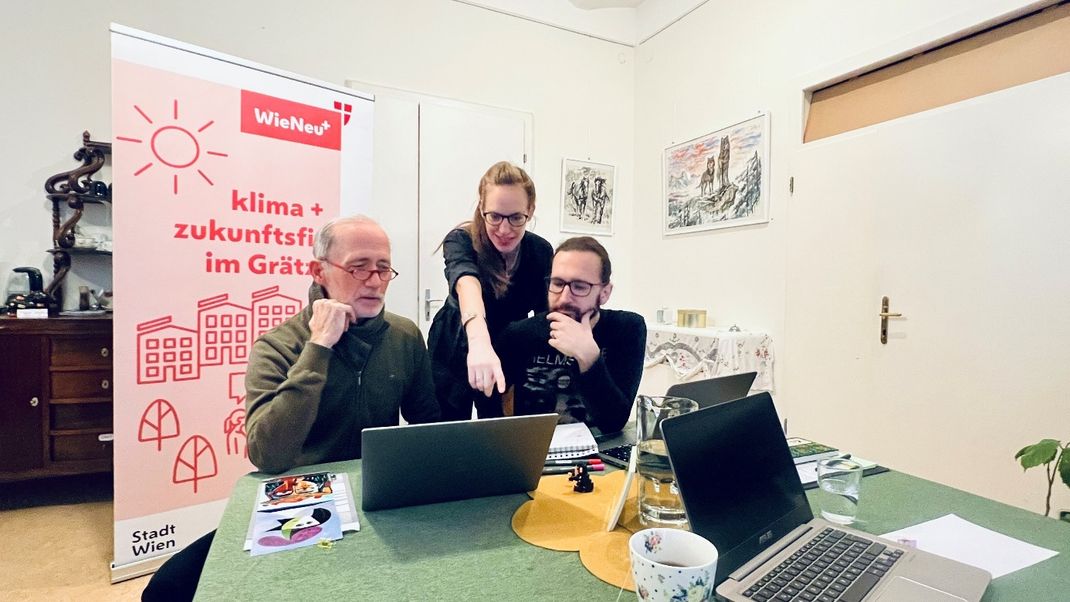 Three people are sitting in a room in front of a laptop; in the centre, a young woman is pointing at the screen. To the left is an elderly gentleman; to the right is a younger man, holding his chin. Behind them is a roll-up banner bearing the WieNeu+ logo.