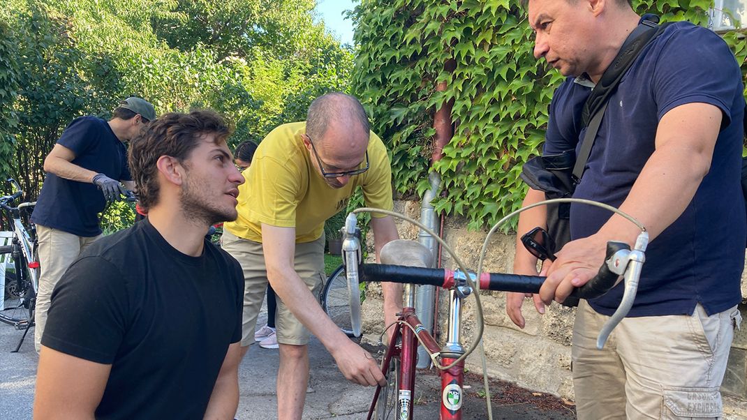 In the centre is an older bicycle, held on the right by a middle-aged man in a T-shirt and shorts. On the left, a younger man in work trousers is crouching, looking up at the man on the right. In the second row, a man in a yellow shirt has his hand resting on the rear of the bicycle. In the background, another man is wearing work gloves. The house wall behind him is covered in ivy, with trees in the far background.