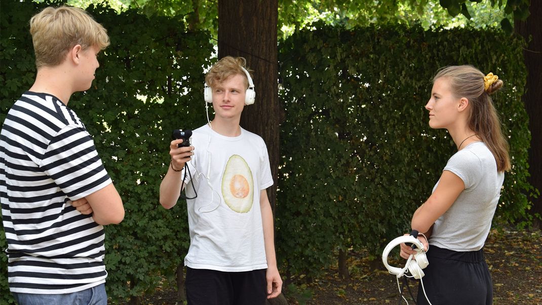 Three teenagers are chatting in the park. The one in the middle is holding the microphone for the one on the right; the girl on the right is listening and holding a pair of headphones in her hands.