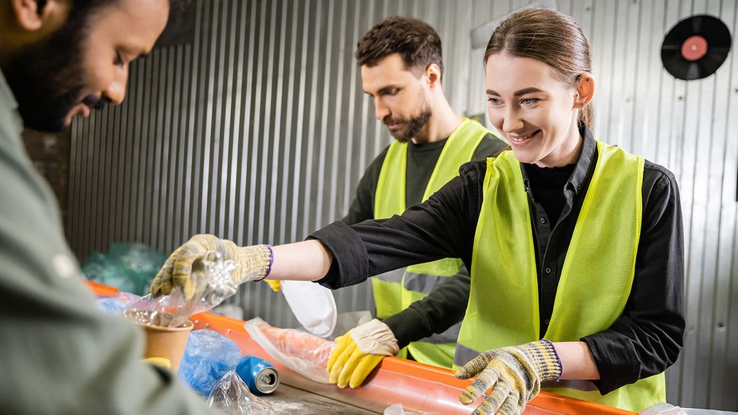 Three people, two of whom are wearing yellow high-visibility vests and gloves, are working head-on at a recycling plant, sorting waste materials on a conveyor belt.