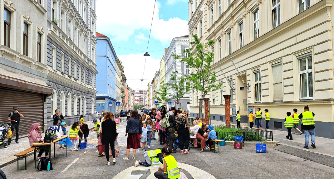 A photo of people on the street with buildings from the Gründerzeit. Some are wearing yellow high-visibility vests, others are playing on the tarmac; on the left, a woman is sitting at a folding table.