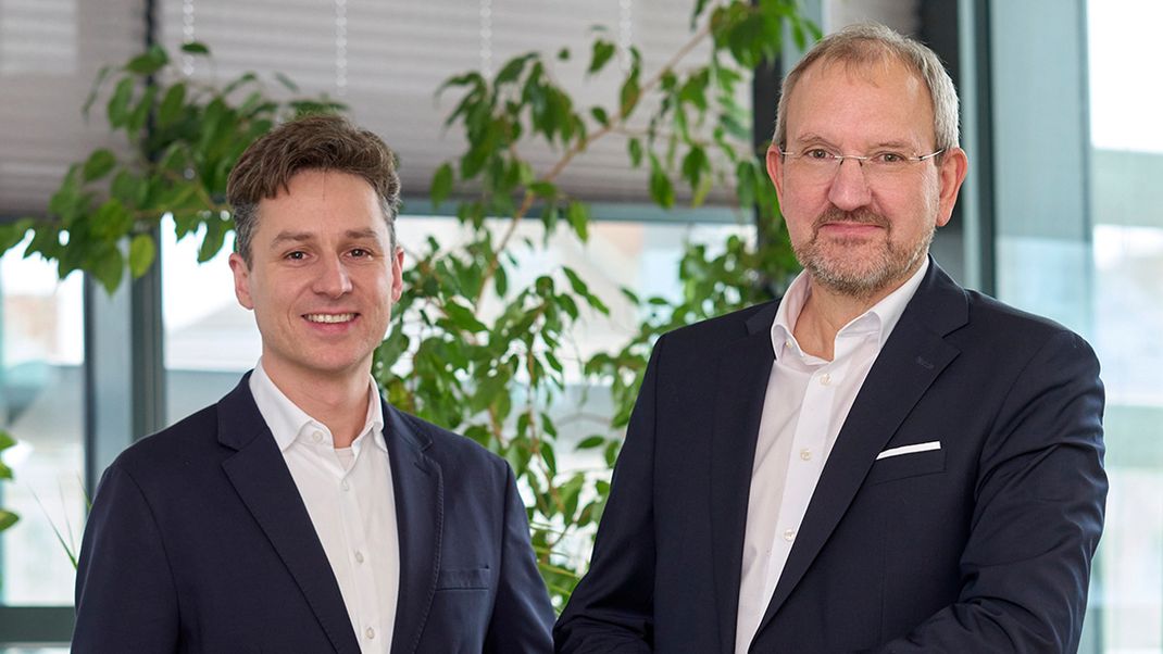 A joint portrait photograph of Stephan Hartmann (left) and Otto Eckl (right), both wearing dark jackets and white shirts and smiling at the camera.