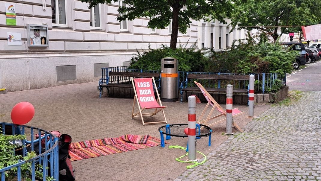 Photo: a rest area on the pavement between two trees, or rather raised tree pits. Two fabric folding chairs, a picnic blanket and a balloon invite you to take a break.