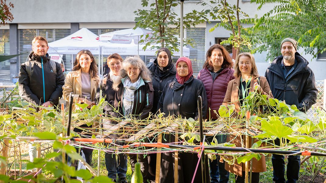 A group of people, mostly women, with a man on either side, are looking at the camera and smiling. The third person from the right is BV Christine Dubravac-Wittholm. In the foreground are green climbing plants on a trellis; in the background is a modern council block.