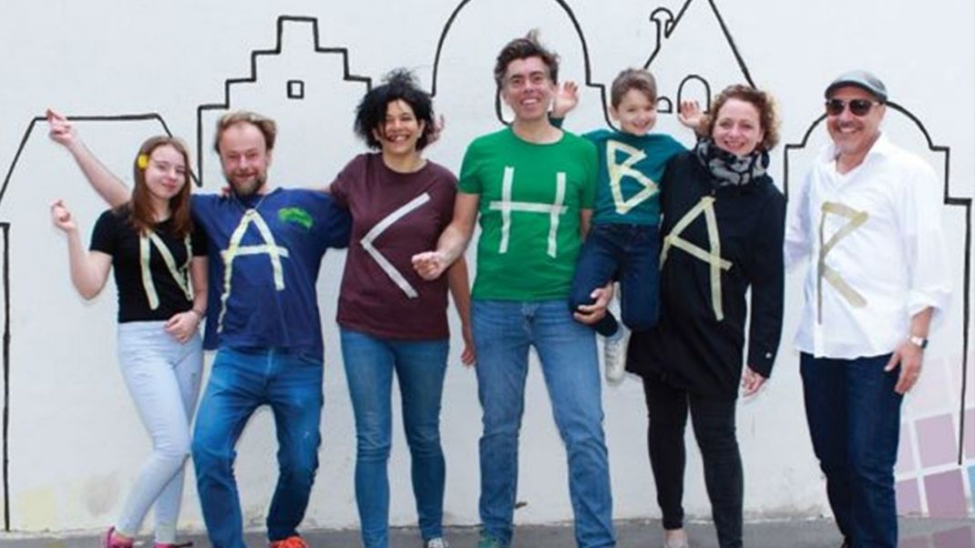 Seven people smiling cheerfully at the camera, wearing T-shirts in different colours that together spell out the word ‘Neighbour’. In the background, against a white wall, is a graphic cityscape with rooftops.