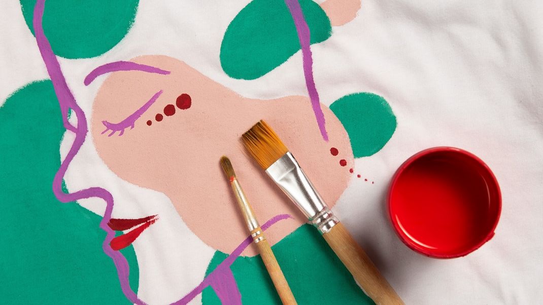 A graphic illustration of a face with purple outlines, containing two paintbrushes and, on the right, a red paint pot, viewed from above.