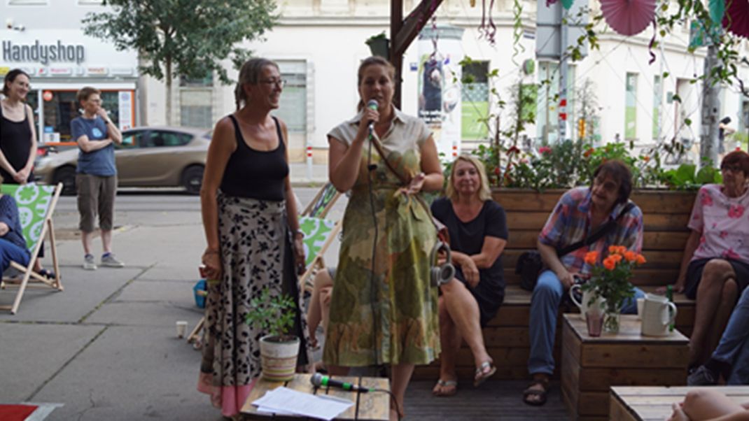 The photo shows people in a neighbourhood oasis within a public urban space. In the centre are two women standing; the one on the right is holding a microphone. 
