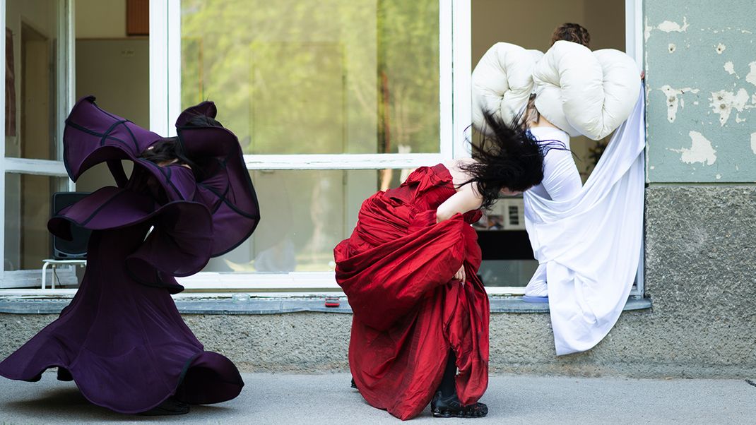 Three female figures in flowing dresses in shades of lilac, crimson and white, dancing in front of a large window. 