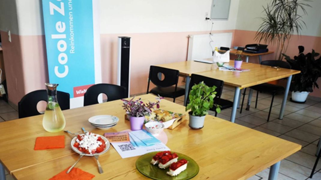 A room with a tiled floor, functional tables and chairs; on one table there are cakes and lemonade laid out; in the background is a roll-up banner bearing the words ‘Cool Zone’