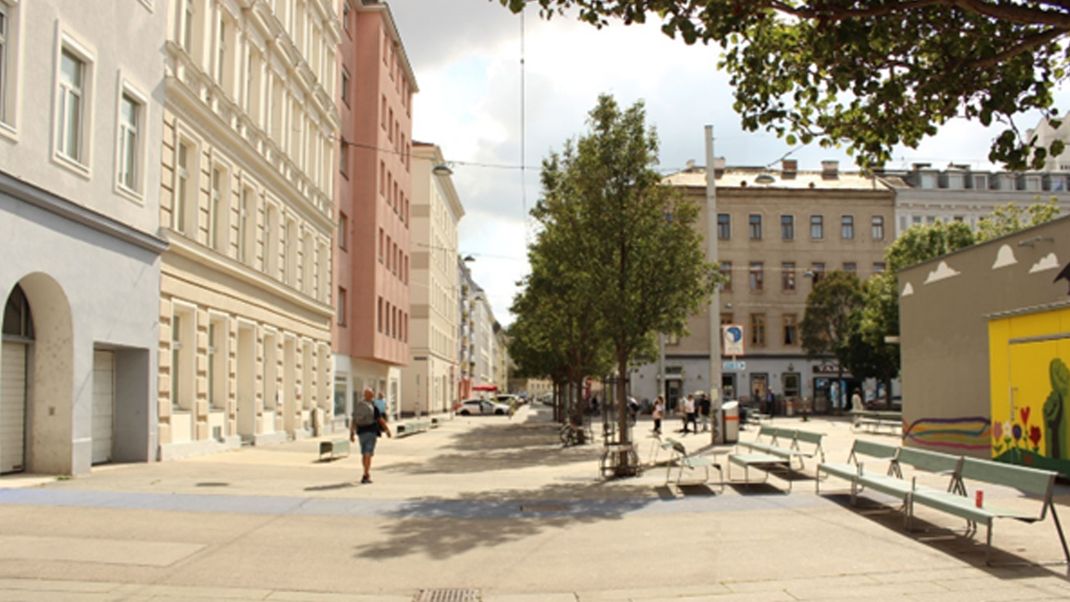 An urban space with old buildings on the left, a brightly paved open area in the centre, and trees and benches in the centre and on the right.