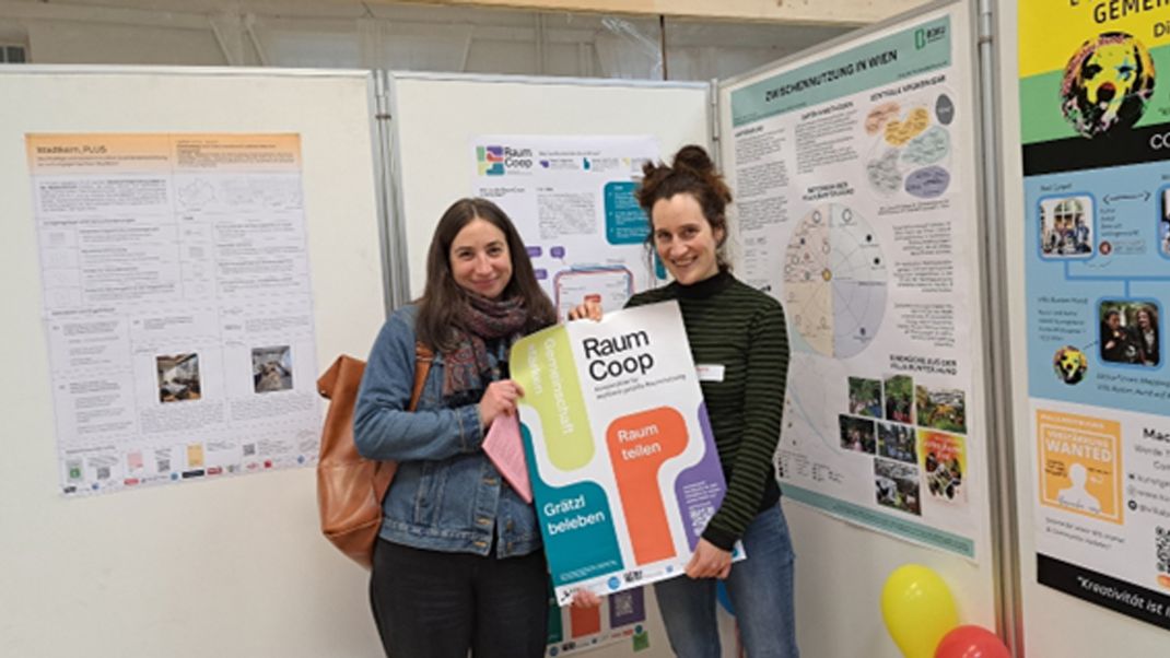 Zwei junge Frauen mit Poster "Raum Coop" gemeinsam in den Händen haltend vor einer Wand mit beschrifteten Projektplakaten.