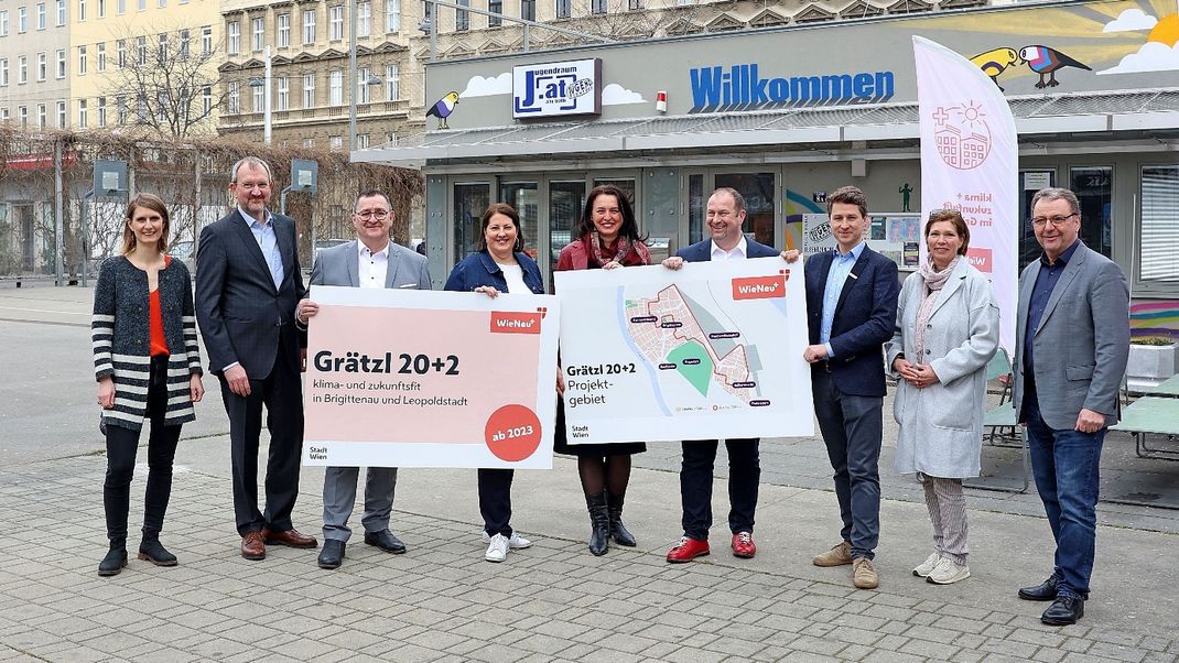 Neun Personen stehen in einer Reihe uns lächeln in die Kamera. Sie halten zwei großformatige Tafeln in der Hand, die auf das Grätzl 20+2 hinweisen sowie als Kartenausschnitt auf dessen geografische Lage. Dahinter ein niedriges Gebäude mit der Aufschrift "Willkommen" sowie Gründerzeitliche Häuser.
