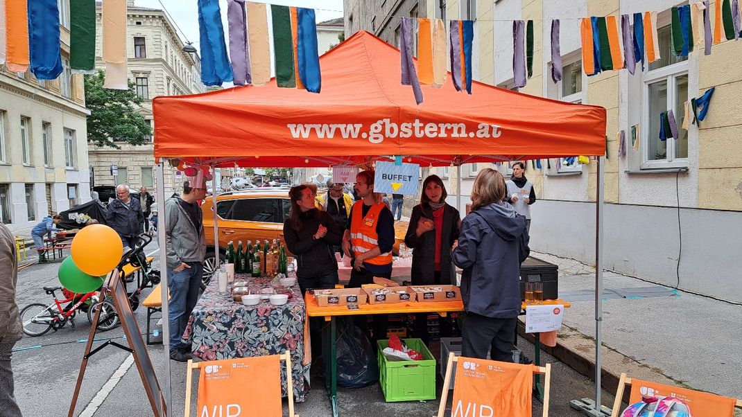 Foto einer Veranstaltung im Straßenraum mit orangem Festzeltdach mit der Aufschrift www-gbstern.at sowie Menschen darunter, die sich unterhalten.