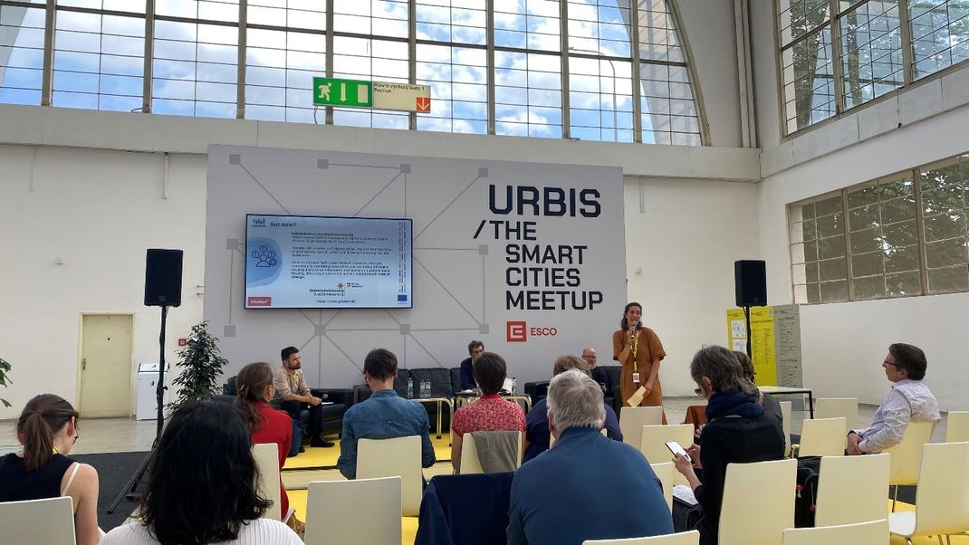 Tagungsfoto mit Helene Scheller auf der Bühne vor Zuhörerinnen unter dem Konferenzplakat "URBIS / The smart Cities Meetup".
