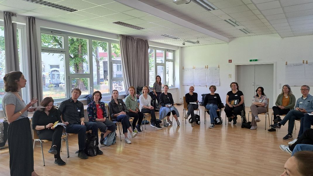 Workshopsituation in einem Raum mit Personen im Halbreis sitzend, vorne rechts Helene Scheller zur Gruppe gerichtet.