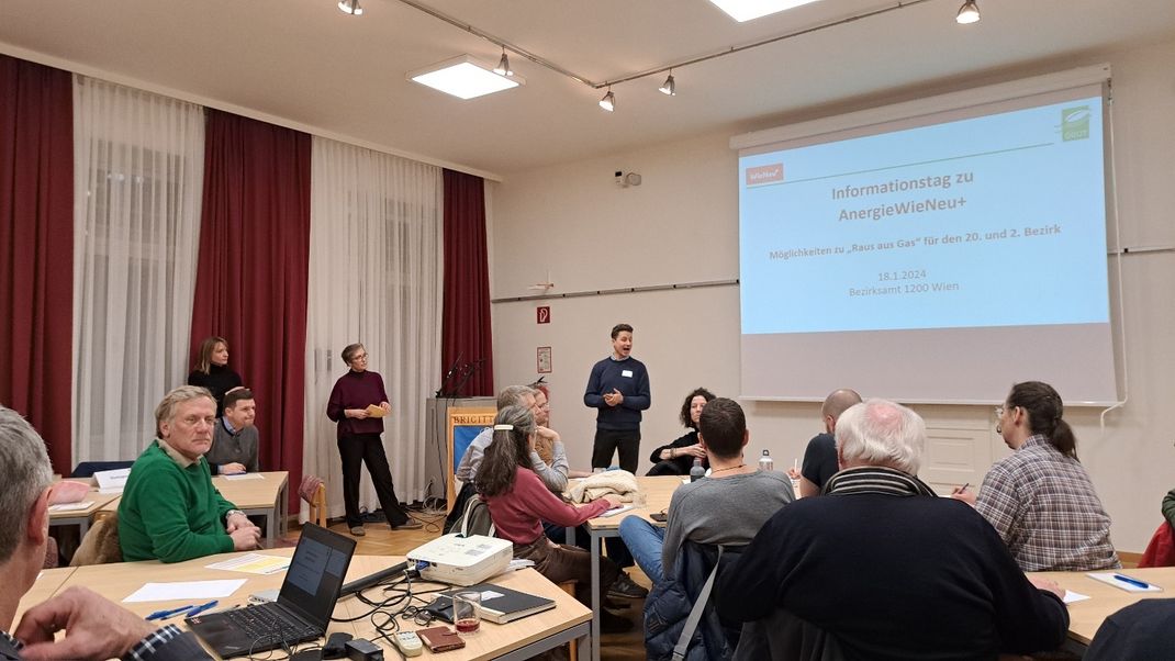 An die 15 Personen sind in einem Vortragssaal versammelt, sitzen an Tischen und hören einem Vortragenden zu, der link vor einer Projektionswand steht.