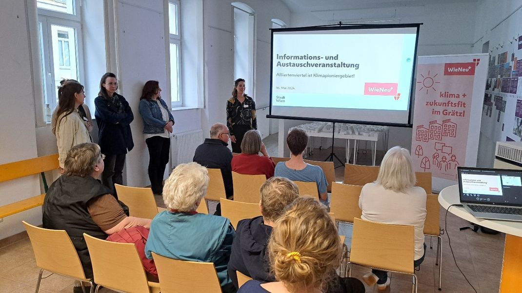 Informationsveranstaltung mit großer Projektionswand im Hintergrund, davor sitzend Personen. Einige stehen am Rande.