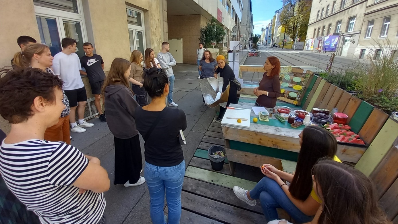 ClimateLibrary

Young people are standing in a semicircle in front of a parklet, listening to a presenter; a table with various utensils is put up on the street.  