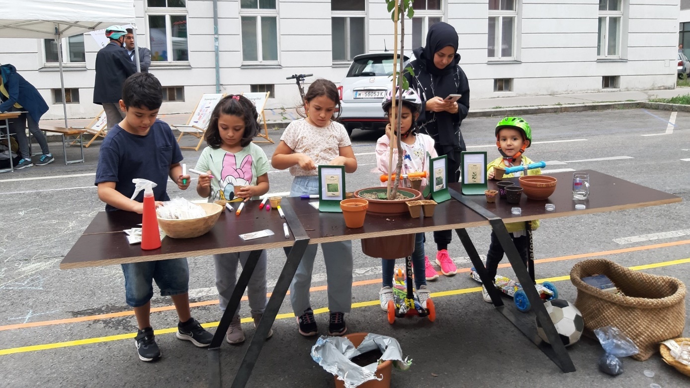 Fünf Kinder beim Pflanztisch mitten im Straßenraum beim Beschriften und Pflanzen mit einer Begleitperson. Copyright: Festival der Ruhe / Florent Souly
