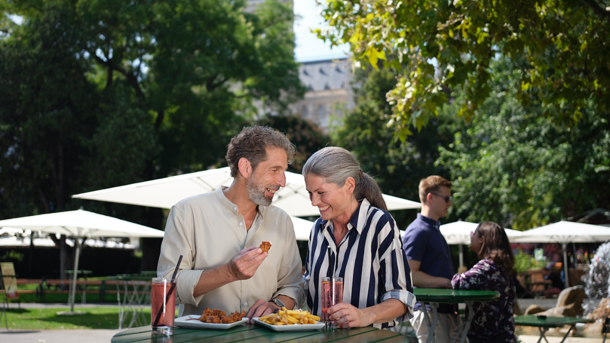 Kapitelbild: A couple is standing at a high table at the Wienliebe Festival, each holding drinks and food. In the background, more high tables and sun umbrellas are visible.