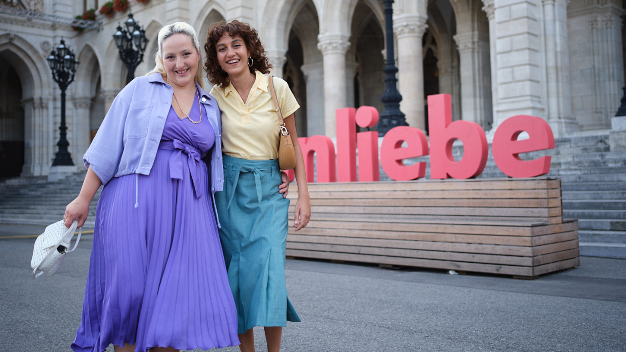 Kapitelbild: Image showing two women standing in front of large red letters spelling “Wienliebe.” In the background, the arches of Vienna City Hall are visible. The scene conveys a friendly and atmospheric mood.