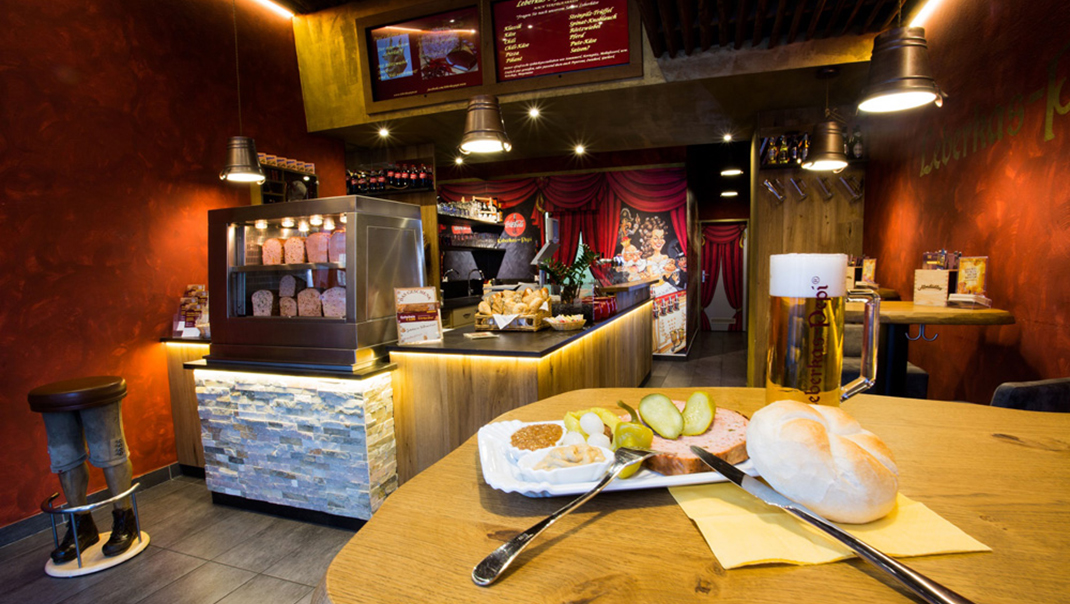 Innenraum einer Leberkas Pepi-Filiale mit einer Vitrine mit Leberkässorten, im Vordergrund ein angerichter Leberkäse mit Semmerl ujnd Beilagen