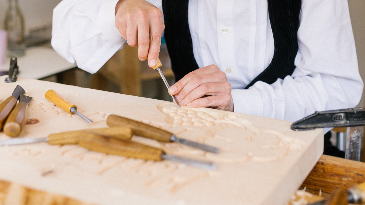Kapitelbild: Fotografie zeigt eine*nHandwerker*in bei Holzschnitzerei, der mit Meißel feine Details in Holzplatte arbeitet. Werkzeuge wie Meißel und Schraubenzieher liegen auf Werkbank, Fokus auf präziser Handarbeit und traditionellem Handwerk.