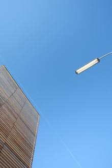 Blick nach oben auf blauen Himmel, links Ausschnitt einer Holzwand, rechts Untersicht auf längliche Straßenlampe.