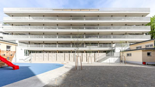 Modernes mehrstckiges Schulgebude mit Balkonen mit Blick auf einen Spielplatz mit weichem Boden und Spielgerten in einem sonnigen, offenen Innenhof.