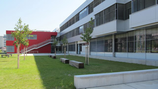 seitliche Ansicht eines langgestreckten Gebäudes mit Fensterbändern, davor Grün; im Hintergrund roter querliegender Trakt