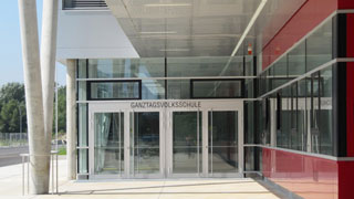 gedeckter Eingang der Ganztagsvolksschule, links vorgelagerte V-Stützenkonstruktion, rechts rote Fassade