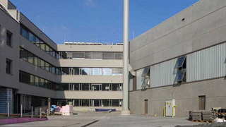 Foto der Hoffassaden und Schornstein. Die Fassade des Büroebäudes sind mit Fensterbändern gegliedert. Die Prüfhalle weist eine fast geschlossene, graue Fassade auf.