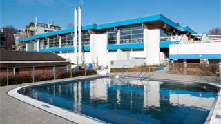 flaches horizontal gegliedertes Gebäude, in Blau, Weiß und Glas, davor ein Schwimmbecken im Außenbereich