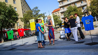 öffentlicher Platz, Menschen diskutieren vor Tafeln und Kleiderpuppen, die T-Shirts mit Kurztexten tragen