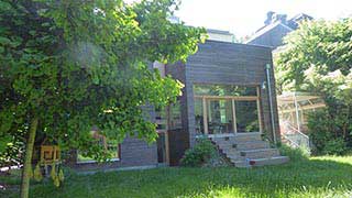 Gartenansicht auf hölzernen Zubau; linker Teil mit zwei übereinanderliegenden Fenstern, rechter Teil mit gläserner Front, zu der Holztreppen führen