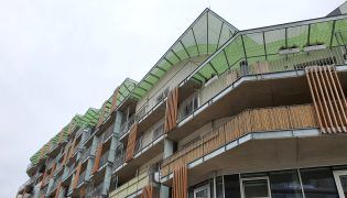 Seitenansicht auf Gebäude mit grünen überkragenden Dachelementen und Fassade, die mit vertikalen Holzelementen gestaltet ist