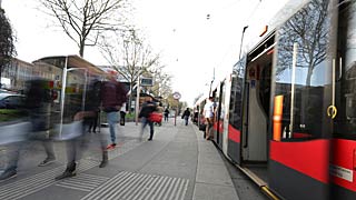 Fotoğrafta bir tramvay var. Aynı zamanda tramvaya binmeye ve inmeye çalışan yolcular gözüküyor.