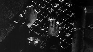 Film still:  Fingers grasping up through a sewer grate