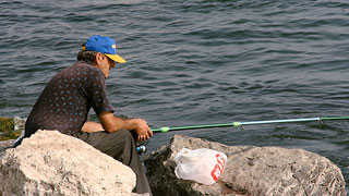 Angler sitzt am Wasser auf einem Stein.