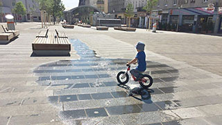 Kleines Kind fhrt mit dem Fahrrad durch eine Wasserlacke der Wiener Wasserwelt